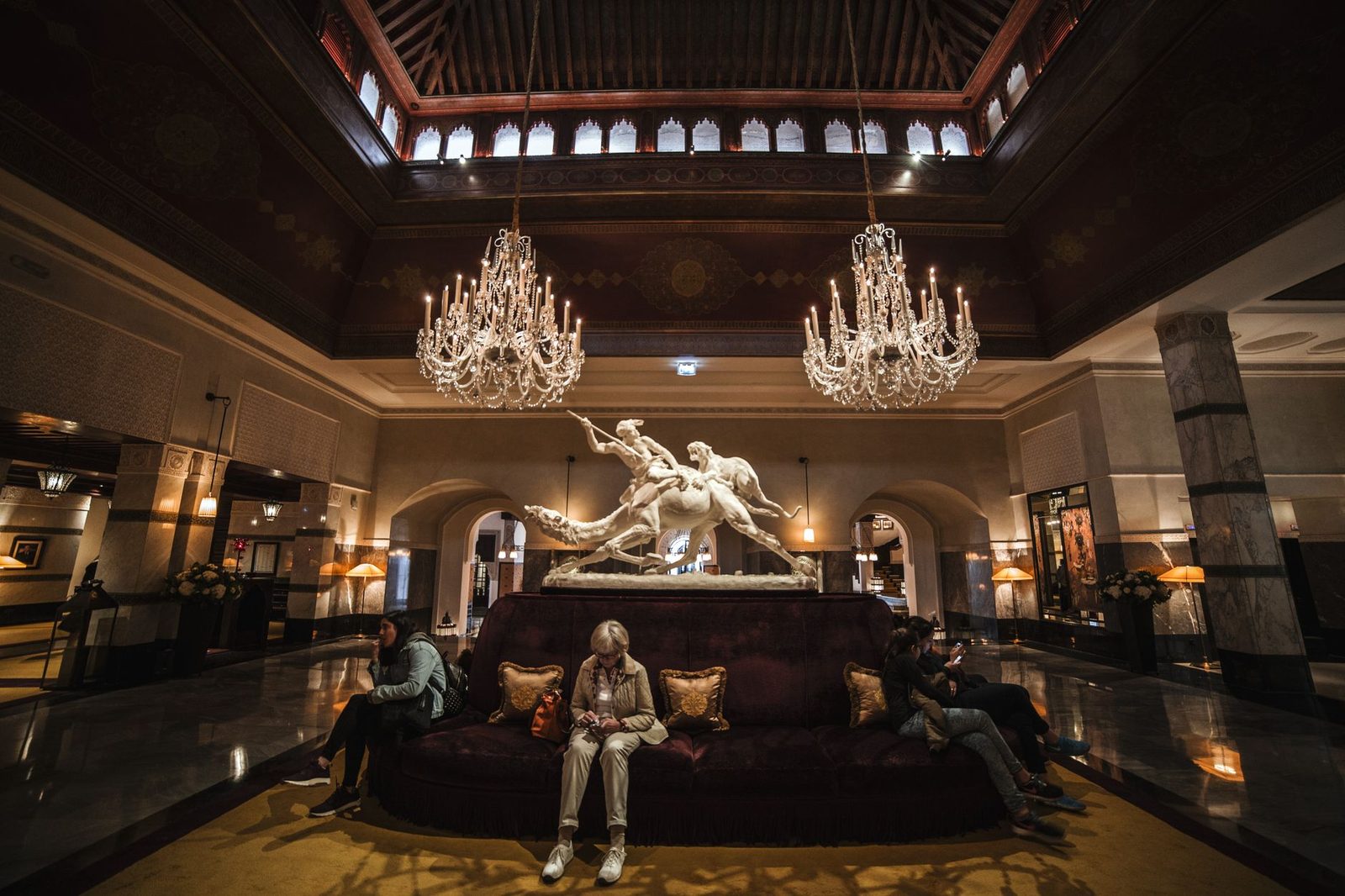 Unrecognizable people sitting on comfortable sofa in spacious hall of luxury hotel with crystal chandeliers and marble s