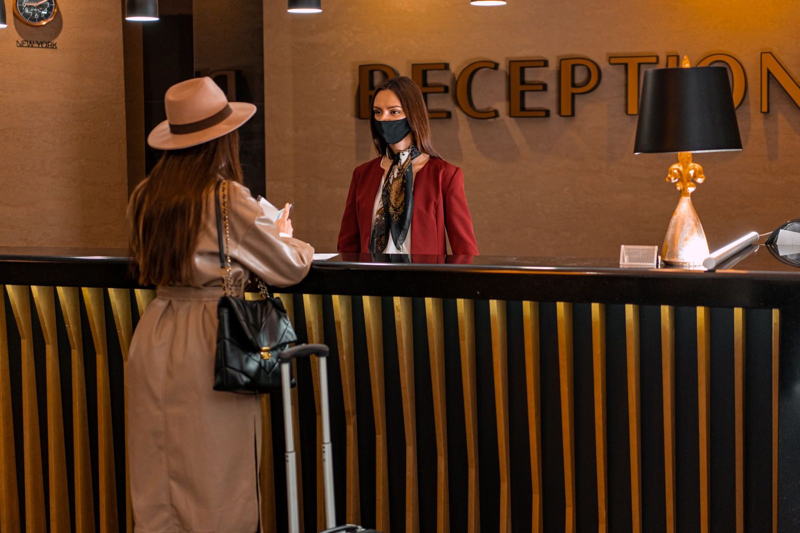 A woman speaks to a receptionist wearing a mask at a hotel reception.