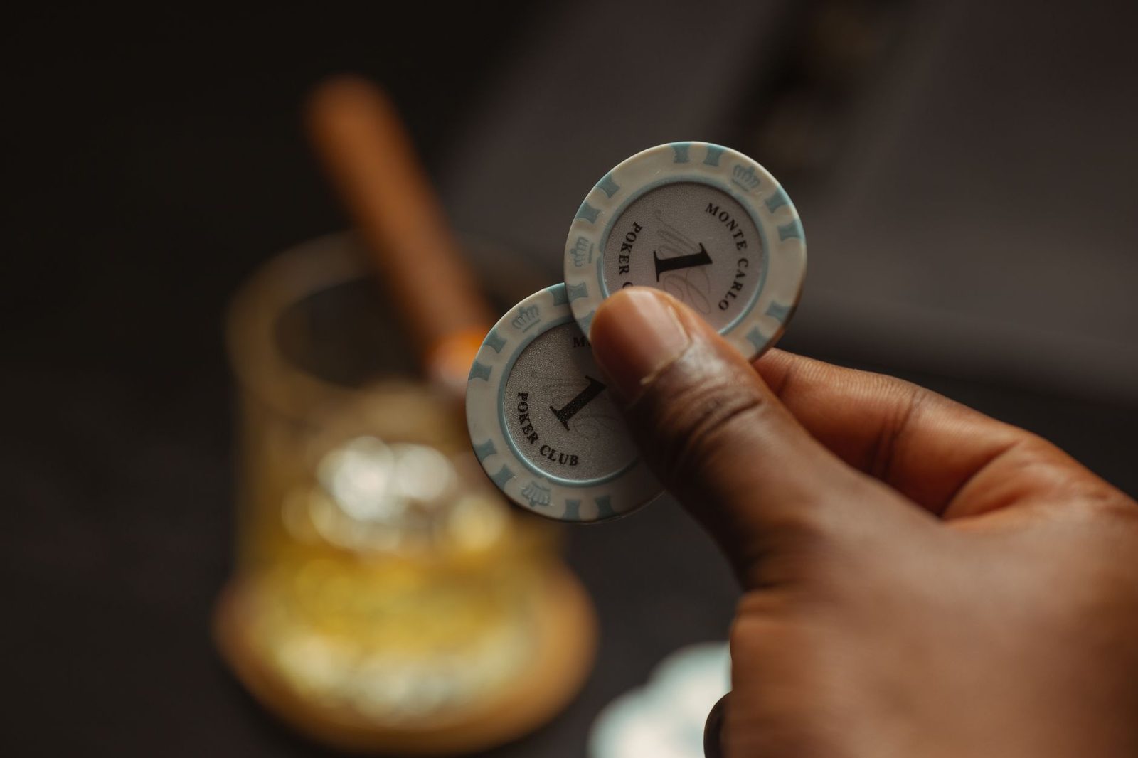 A close-up of a hand holding poker chips with a blurred drink in the background, capturing a casino atmosphere.