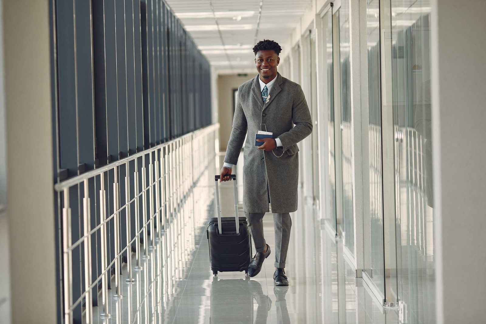 Full length of happy African American male in stylish classy gray clothes with suitcase and passport walking in contempo