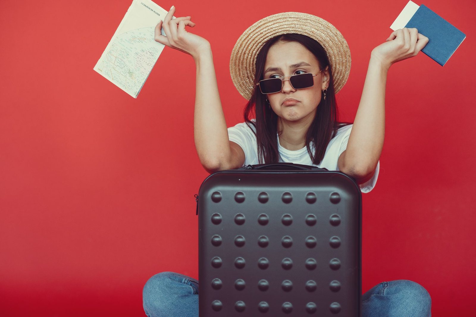 Doubters young stylish woman in wicker hat and trendy sunglasses sitting behind black suitcase and shrugging with passpo