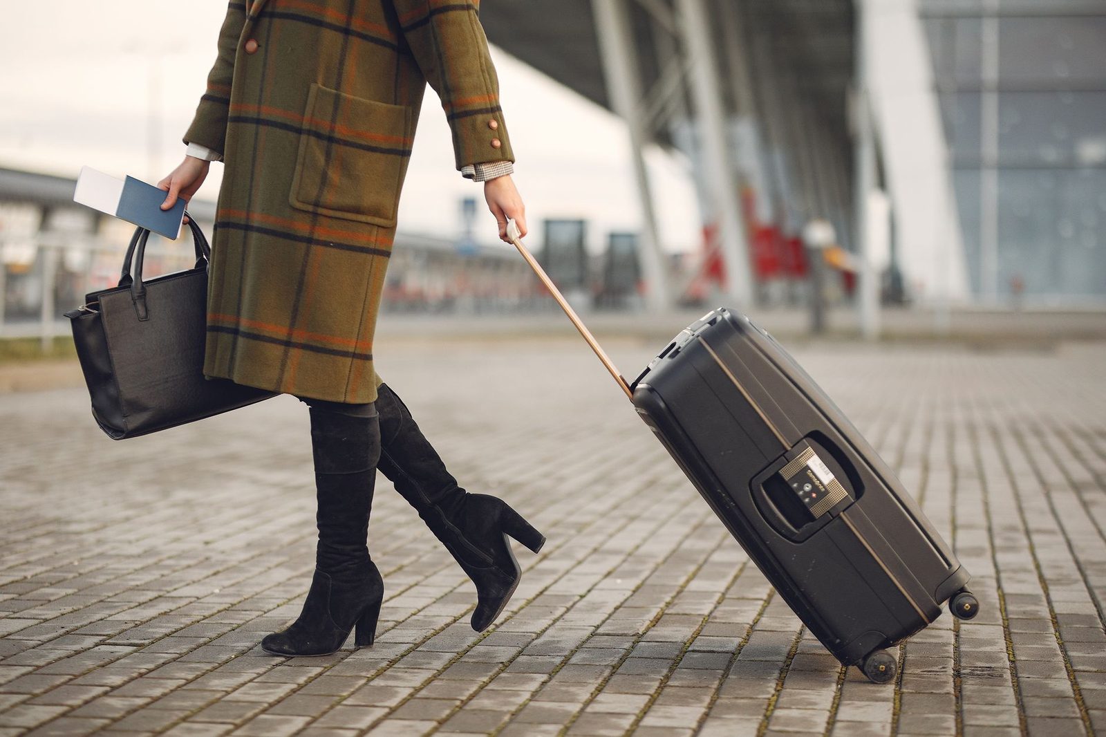 Crop young female traveler in trendy plaid coat and high boots with luggage and documents strolling on pavement near con