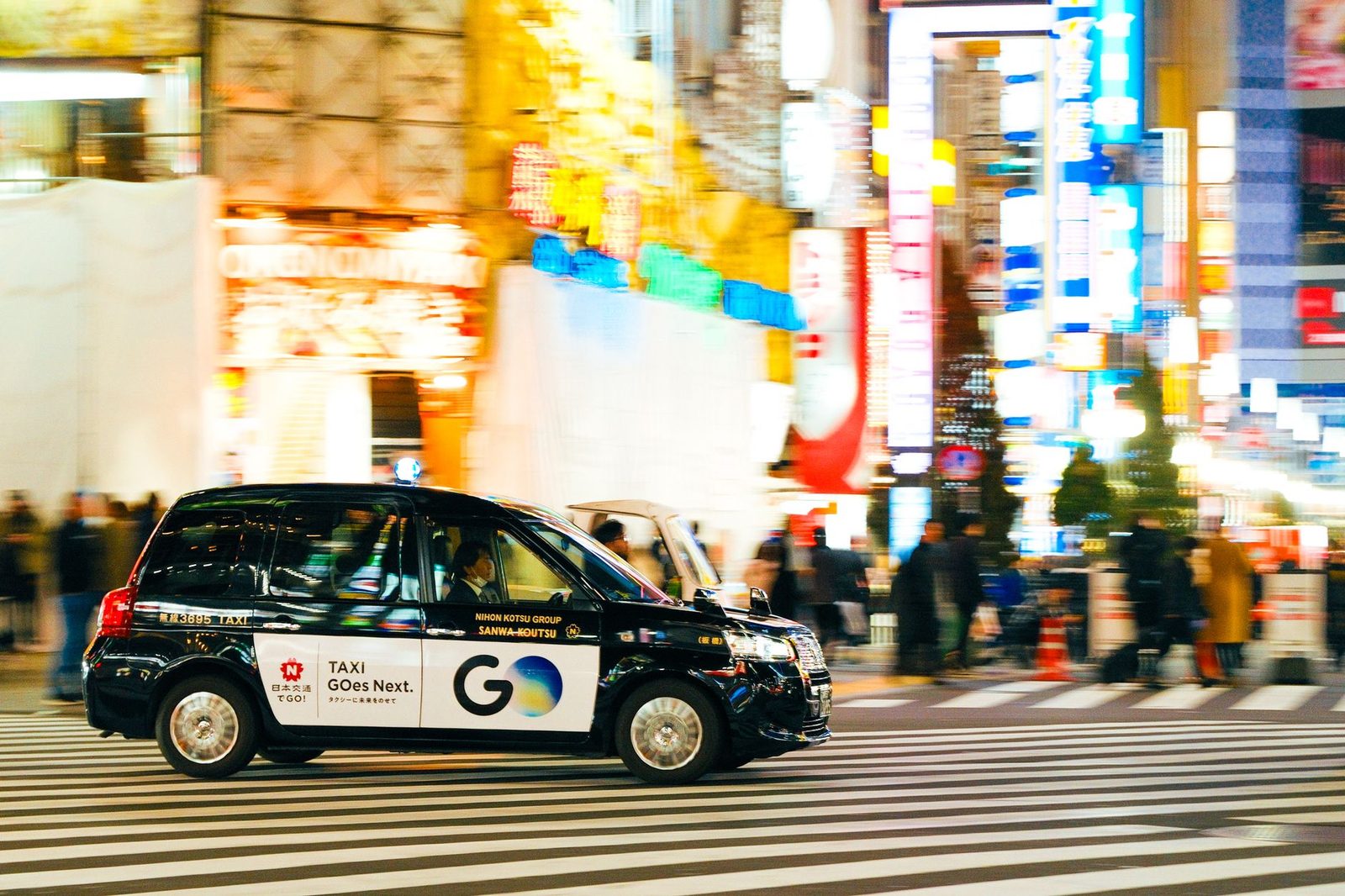 A black taxi driving through a bustling, colorful city street at night under bright neon lights.