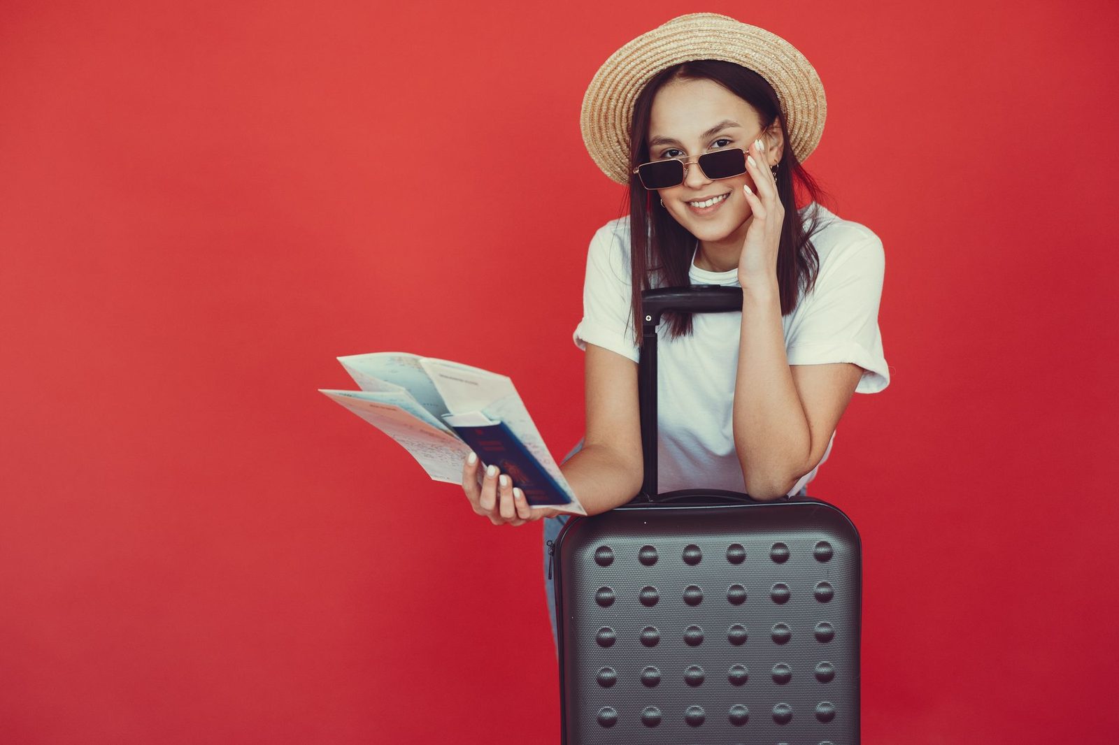 Delighted young woman in straw hat in casual clothes leaning on suitcase and checking tickets before traveling on red ba