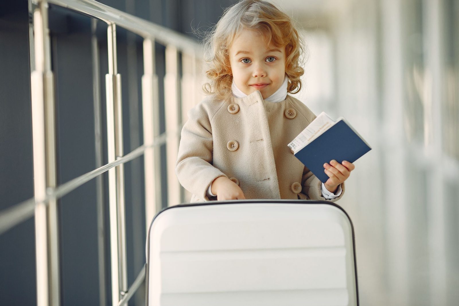 Adorable little girl with blond curly hair in stylish beige coat standing with white suitcase and passport in airport co